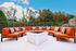 Inviting outdoor patio with vibrant orange sectional seating and a central fire pit, surrounded by lush greenery under a clear blue sky.