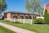 Charming brick apartment complex with private balconies, surrounded by lush greenery and a white picket fence on a sunny day.