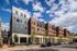 Modern apartment building with a blend of brick and metal facade, lined with trees on a bustling urban street under a vibrant blue sky.