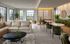 Chic and modern living space with plush beige sofa, sleek black coffee table, and elegant green chairs. Features large windows, abundant natural light, and lush greenery accents.