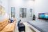 Bright and inviting apartment interior featuring a cozy tan couch, a sleek TV with scenic views, and a modern bed, all complemented by large windows with natural light.