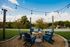 Charming outdoor seating area with blue Adirondack chairs around a rustic fire pit, under glowing string lights, surrounded by lush greenery and a scenic countryside view.