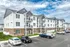 Modern apartment complex with elegant architecture featuring white siding and brick accents, nestled in a serene setting with ample parking and bright blue skies.