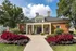 Charming brick apartment building framed by lush landscaping, vibrant flowers, and mature trees under a bright blue sky.