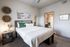 Inviting bedroom with elegant decor, featuring a cozy bed with white and green accents, stylish wall art, and a glimpse into a modern, plant-filled bathroom.