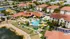 A stunning aerial view of a vibrant apartment complex featuring a large, resort-style swimming pool surrounded by lush palm trees and elegant red-tiled roofs, nestled in a picturesque landscape.