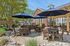 Charming outdoor patio with shaded seating, lush greenery, and a built-in BBQ area under a bright blue sky.