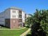Charming four-story apartment building with balconies, surrounded by lush greenery and a winding pathway, under a clear blue sky.