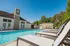 Luxurious outdoor pool area with cozy sun loungers and elegant clubhouse, surrounded by lush greenery under a clear blue sky.