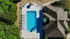 Bird's-eye view of a luxurious outdoor pool area with stylish lounge chairs and a shaded seating spot, adjacent to a modern building and surrounded by lush greenery.