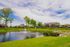 Charming countryside homes overlooking a serene pond with a feature fountain, lush greenery, and vibrant sky.