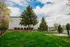 Spacious green courtyard with lush grass, tall trees, and a serene bench area, surrounded by a secure fence under a bright blue sky.