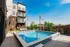 Modern apartment building with balconies overlooking a sparkling pool, surrounded by lush greenery and lounge chairs under a clear blue sky.