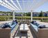 Chic outdoor seating area with wicker furniture and vibrant cushions under a stylish pergola overlooking serene pond and modern townhouses.