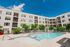 Modern apartment complex with a sparkling swimming pool, sun loungers, and lush greenery under a bright blue sky.