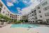 Modern apartment complex with a sunlit courtyard, featuring a sparkling swimming pool and lush greenery under a clear blue sky.