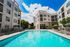 Modern apartment complex with a luxurious swimming pool, surrounded by elegant white buildings and lush greenery, under a bright blue sky.