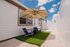 Charming outdoor patio with cozy seating and a large umbrella, set against a modern white exterior with vibrant green artificial turf and stone accents under a bright blue sky.