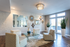 Modern living area with stylish decor, featuring geometric mirrors, plush seating around a glass coffee table, large windows, and vibrant green plants.