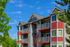Charming three-story apartment building with vibrant red accents, private balconies, and lush greenery under a clear blue sky.