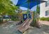 Charming playground with slides and climbing features, shaded by a blue canopy, surrounded by lush greenery and modern residences.