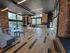 Modern communal space with stylish wooden flooring, exposed brick walls, and ample natural light from large windows, featuring versatile seating options and an open, airy atmosphere.