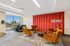 Modern lounge area with vibrant red accent wall, plush orange chairs, and a massage chair, featuring large windows with city view.