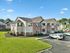 Charming apartment building with brick facade and manicured green lawns, featuring ample parking and serene surroundings under a clear blue sky.