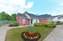 Charming red brick apartment building with a welcoming entrance, surrounded by manicured greenery and vibrant flower beds under a clear blue sky.