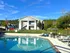 Charming apartment complex with modern architecture, set against a lush green backdrop, featuring a sparkling outdoor pool and comfortable poolside seating under a bright blue sky.