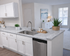 Modern kitchen with sleek marble countertops, white cabinetry, and integrated stainless steel dishwasher, opening into a bright living space with large windows.