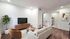 Chic modern living room with cozy beige sofa, stylish wood furniture, and green plant, opening into a sleek white kitchen.
