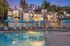 Luxurious apartment community with elegant architecture, featuring a pristine pool surrounded by lounge chairs and umbrellas, set amidst lush palm trees during a serene twilight.