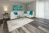 Elegant living room with a plush white sectional sofa, teal accents, modern glass tables, and natural light streaming through vertical blinds.
