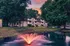 Charming apartment complex nestled among lush trees, featuring a serene pond with a glowing fountain at sunset.