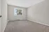 Bright and spacious empty room with soft carpet flooring, neutral gray walls, and a large sunny window offering a view of lush greenery outside.