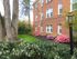 Charming brick apartment building with vibrant garden, featuring lush greenery, blooming flowers, and a quaint lamp post in a serene residential setting.