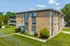 Charming brick apartment building surrounded by lush green lawn, offering sunlit windows and a welcoming entrance in a peaceful neighborhood.