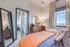 Chic urban bedroom with modern decor, featuring a cozy bed with orange linens, stylish dresser with a round mirror, and large windows offering breathtaking city views.