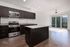Modern kitchen with sleek dark cabinets, stainless steel appliances, and a spacious granite island, opening into a sunlit living area with large windows.