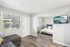 Bright and modern living area with a sectional sofa, wall-mounted TV, and a glimpse into a cozy bedroom; large windows offer a view of the serene outdoor space.