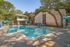 Inviting apartment complex with a sparkling pool, surrounded by lush trees and charming stone buildings, offering a serene outdoor retreat.
