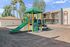 Vibrant playground area with colorful slides and climbing equipment beside modern apartments, complemented by tall palm trees and a clear blue sky.