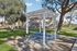 Inviting community picnic area with a pergola, blue picnic table, and barbecue grill, set amidst lush green gardens and charming residential buildings.