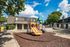Charming apartment community with vibrant playground featuring slides and climbing structures, surrounded by lush trees and modern buildings under a clear blue sky.