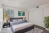 Elegant bedroom with modern decor, featuring a plush bed set against a serene artwork, natural light streaming through large windows, and a cozy ambiance enhanced by a stylish rug and vibrant green plant.