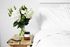 bed with white covers and wooden side table with plants in a vase
