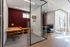 Modern office space with glass walls, featuring a stylish red-accented private office and a cozy seating area, illuminated by natural light.