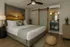 Inviting bedroom with sleek wooden floors, a cozy bed adorned with plush pillows and a soft throw, framed by a sunset mural. Features include stylish plant decor and a modern sliding barn door leading to an elegant vanity area.