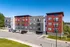 Contemporary apartment building with vibrant red and gray exterior, surrounded by lush greenery and ample parking space.